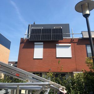 De foto toont een installatie van zonnepanelen op een woning, bevestigd aan het dak met een heldere blauwe lucht op de achtergrond.
