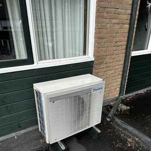 Een buitenunit van een airco-systeem, geplaatst naast een muur met zichtbare leidingen en kabels.