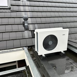 Deze installatie toont een buitenunit van een warmtepomp, geplaatst op een plat dak met enkele begroeiing en nabij raamdecoratie.
