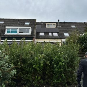 De afbeelding toont een woning met zonnepanelen op het dak, omgeven door een groene tuin en een bewoner die langsloopt.