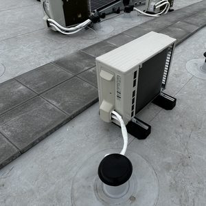 De afbeelding toont een airconditioninginstallatie met de buitenunit op een dak, met leidingen en ondersteuningen zichtbaar.