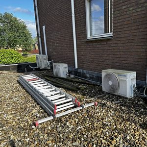 De afbeelding toont een buitenunit van een airconditioninginstallatie, geplaatst op een plat dak met een ladder en grind eromheen.