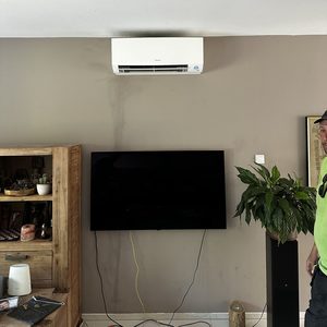 Een wandairconditioner is geïnstalleerd, met zichtbare leidingen langs de muur en een televisie op de achtergrond.