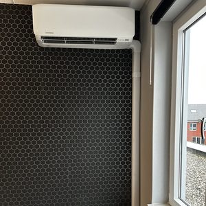 Een wand-airco-unit is geïnstalleerd aan een hexagonale wand, met zichtbare leidingen en een stopcontact eronder.