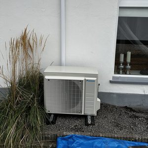 De afbeelding toont een buitenunit van een airconditioning systeem, bevestigd aan de muur, met leidingen die naar binnen lopen.