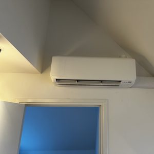 Een wandairco-installatie bevestigd aan een schuin plafond, met zichtbare luchtkanalen en een deur op de achtergrond.