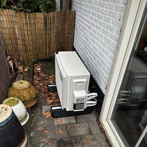 De afbeelding toont een airco-installatie buiten, geplaatst op een bak met leidingen aangesloten voor koeling en verwarming.