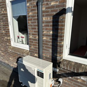 Een buitenunit van een airconditioningsysteem, geïnstalleerd aan de muur van een gebouw, met aansluitingen voor stroom en koelleidingen.
