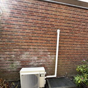 Een buitenunit van een airco-installatie, geplaatst tegen een bakstenen muur met leidingen en een rooster voor luchtcirculatie.
