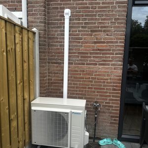 Een airconditioningseenheid van Daikin is geïnstalleerd aan de buitenwand van een woning, met de afvoerpijp zichtbaar langs de muur.