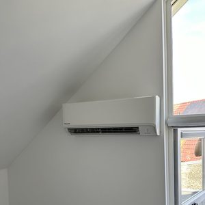 Een wandmodel airconditioningseenheid geïnstalleerd in een kamer met schuin plafond, naast een geopend raam.