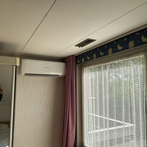 Een muurgeïnstalleerde airconditioner in een slaapkamer, met een spiegel en een raam op de achtergrond.