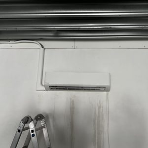 Een wandmontage airconditioner afgesloten met leidingen, zichtbaar op een vlakke muur. Er is een ladder in de hoek zichtbaar, wat wijst op een recent installatieproces.