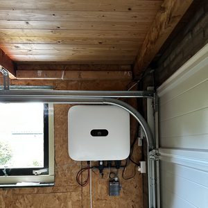De afbeelding toont de omvormer van een zonnepaneleninstallatie, gemonteerd in een garage. De bekabeling en componenten zijn zichtbaar.