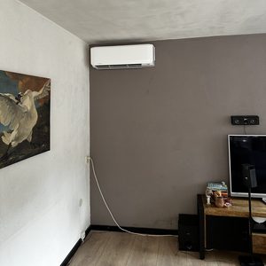 Een luchtgekoelde airco-installatie bevestigd aan de muur in een woonkamer, met zichtbare kabels en meubels in de nabijheid.