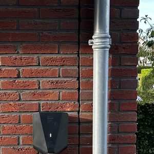 De afbeelding toont een laadpaal die aan een muur is bevestigd, met zichtbare kabels en een metalen pijp, wat duidt op een installatie voor elektrische voertuigen.