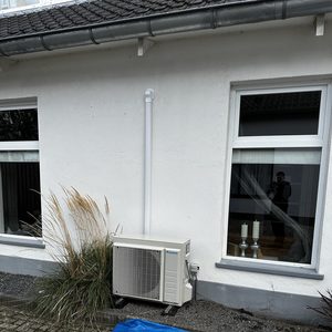 De afbeelding toont een buitenunit van een airconditioning installatie, geplaatst tegen een muur, met leidingen en een geordende omgeving.