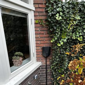 Een laadpaal is geïnstalleerd naast de buitenmuur, omgeven door planten. De installatie is net en discreet.