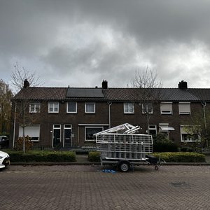 De afbeelding toont een woonhuis met zonnepanelen op het dak, met een aanhangwagen vol materialen ernaast, wat wijst op een installatieproject.