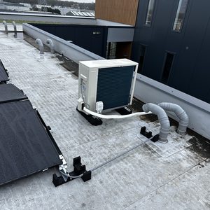 Een airconditioning installatie op een plat dak, met zonnepanelen op de achtergrond. Dit systeem is bedoeld voor het koelen van de binnenruimtes.